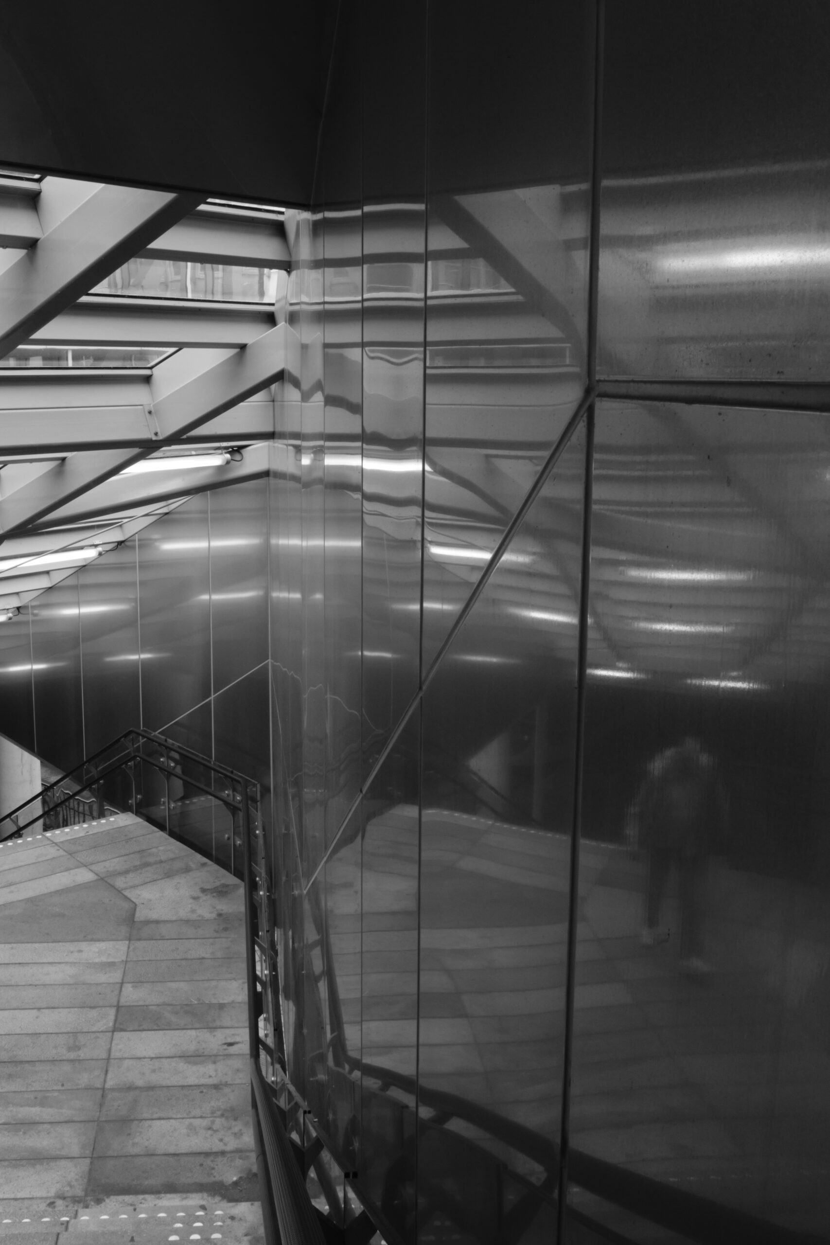 Reflectie van een persoon in een metalen wand waarbij een deel van de trap aan de linkerkant nog zichtbaar is.